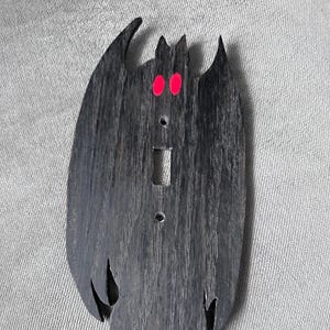 May include: A dark gray wooden light switch cover in the shape of a creature with large wings and red eyes. The cover has a cutout for the switch and screw holes for mounting. The design is a unique, decorative element for any room.