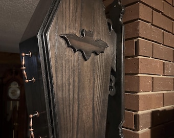 Può includere: Una scatola di legno nera a forma di bara con un disegno di pipistrello sul davanti. La scatola è montata su una parete e ha una staffa di metallo nero.