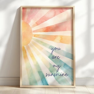 Puede incluir: Una impresión en acuarela enmarcada con un diseño de rayos de sol en tonos naranja, rosa, amarillo, verde y azul. Las palabras "you are my sunshine" están escritas en cursiva. La obra de arte se exhibe en una pared blanca y un suelo de madera.