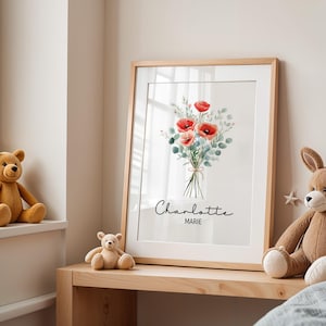 Puede incluir: Una impresi&oacute;n art&iacute;stica enmarcada con un ramo de acuarela de amapolas rojas y follaje verde, atado con hilo. La impresi&oacute;n est&aacute; en un marco de madera clara e incluye los nombres "Charlotte Marie". La obra de arte se exhibe en una estanter&iacute;a de madera.