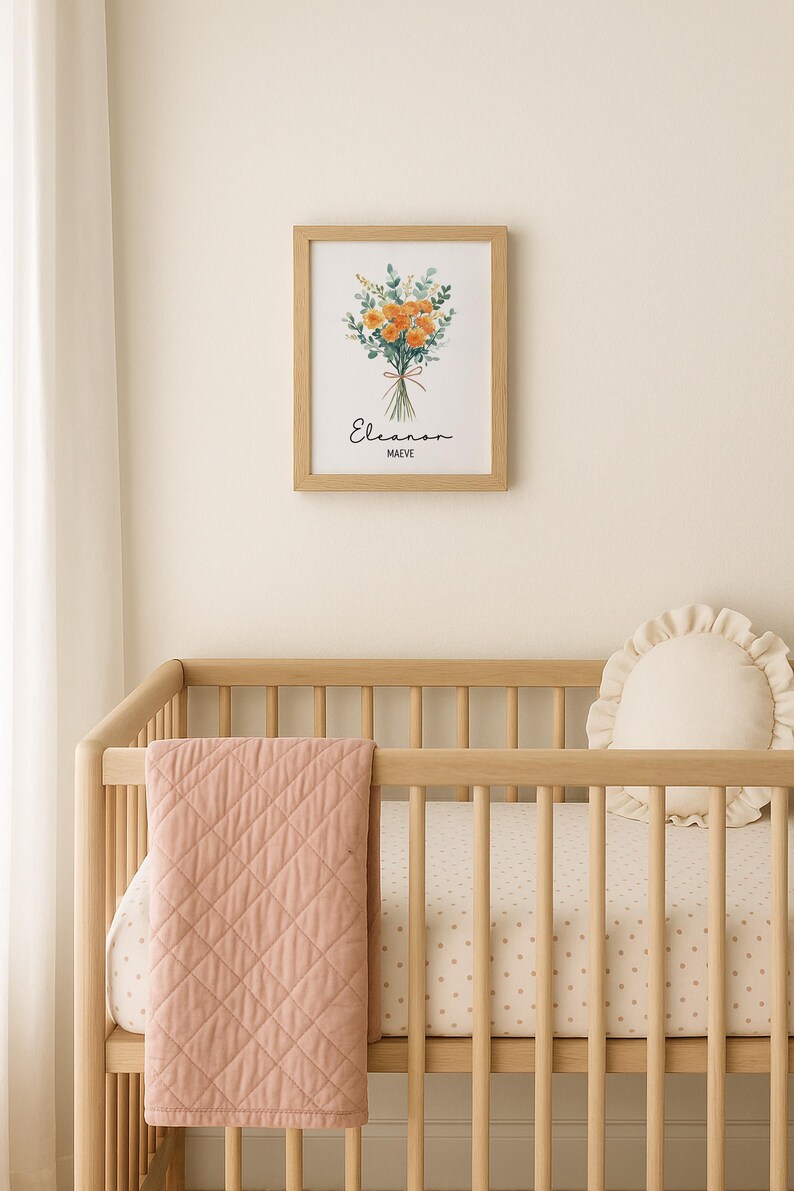 May include: A framed floral print with orange and green flowers hangs above a wooden crib. The print includes the name "Eleanor" and the word "MAEVEE". A pink quilted blanket and a cream-colored pillow with a ruffled edge are in the crib.