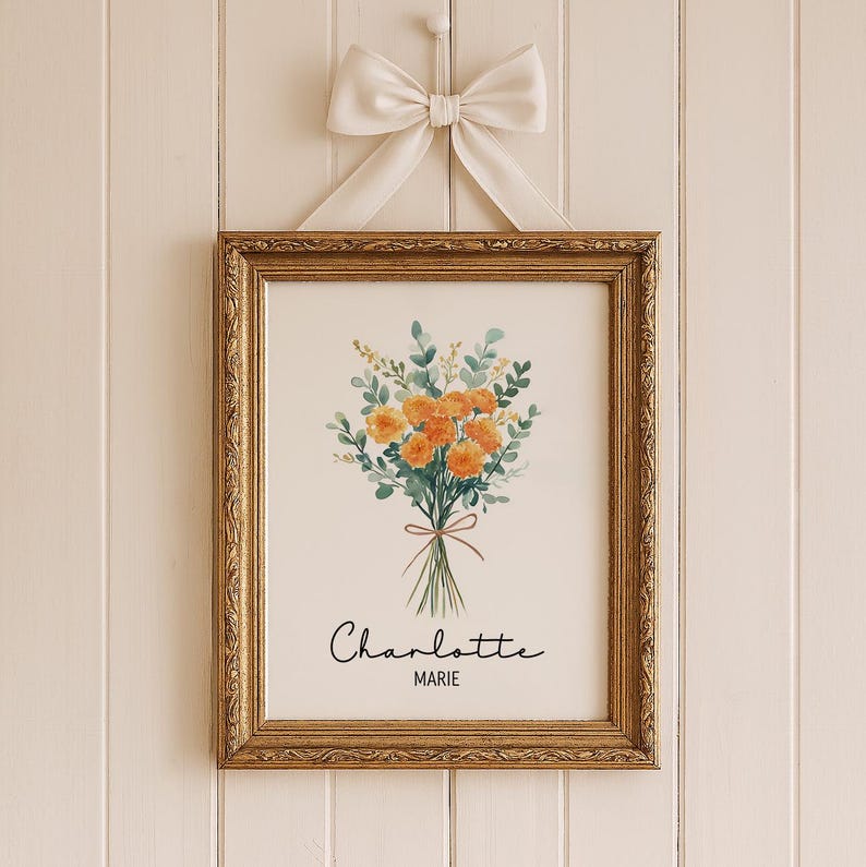 May include: A framed floral print featuring a bouquet of orange flowers and green foliage tied with a brown ribbon. The print is in a gold ornate frame and is hanging from a white bow. The name "Charlotte Marie" is written in script below the flowers.