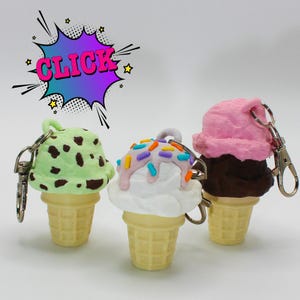 Puede incluir: Tres llaveros de cono de helado. Uno tiene helado verde menta con manchas marrones, otro tiene helado blanco con chispas y el último tiene helado rosa. Todos tienen bases de cono amarillas y llaveros plateados. La palabra "CLICK" está en una burbuja de estilo cómic.