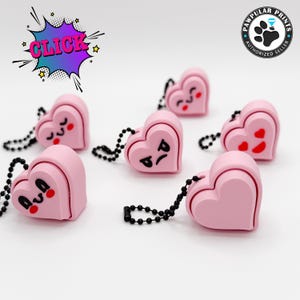 May include: Six pink heart-shaped keychains with different facial expressions. Each keychain has a black chain.