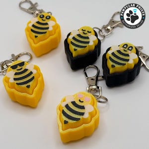May include: A collection of bee-themed keychains. Each keychain features a yellow and black bee design, some with sunglasses or hearts, set in a yellow or black honeycomb-shaped base. Silver-colored clasps are attached.