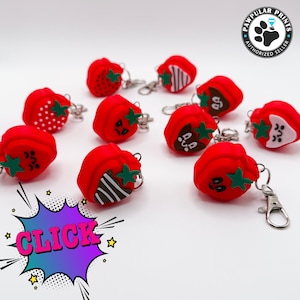 May include: A set of ten red strawberry-shaped keychains with different designs. Each keychain has a silver metal ring and a small green leaf. The keychains are arranged on a white surface. CLICK.
