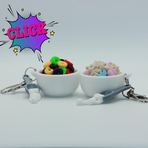 May include: Two white ceramic bowl keychains, each filled with colorful cereal. One bowl has a variety of colorful cereal rings, and the other has cereal with pastel marshmallows. The keychains have a spoon and a silver clasp. The word "CLICK" is in a comic book style bubble.