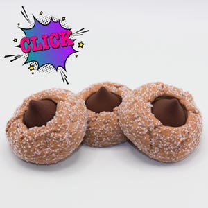 Puede incluir: Tres galletas marrones con un centro de chocolate negro, espolvoreadas con azúcar blanca. Las galletas están dispuestas sobre una superficie blanca. Un bocadillo de estilo cómic con la palabra "CLICK" en rosa y azul está en la esquina superior izquierda.