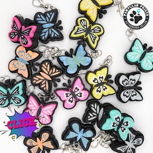 Pode incluir: Uma coleção de chaveiros de borboleta coloridos com fechos de metal prateados. Os chaveiros são feitos de um material macio e flexível e apresentam uma variedade de designs de borboleta. Os chaveiros estão dispostos em uma pilha sobre um fundo branco. O texto "Pawpular Prints" e "Authorized Seller" é visível no canto superior direito da imagem. O texto "Click" é visível no canto inferior esquerdo da imagem.