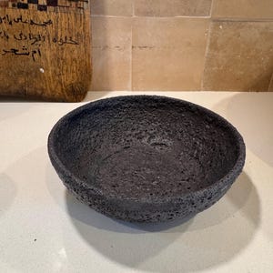Peut inclure: Un bol rond et peu profond d'une couleur noir charbon foncé avec une surface très texturée et poreuse rappelant la roche volcanique. Le bol présente un bord épais et irrégulier ainsi qu'un bassin intérieur profond et cratérisé.