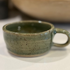 May include: A low-profile ceramic mug with a wide, circular opening and a single side handle. The exterior features a mottled sage green glaze with dark brown speckles and subtle horizontal ridges along the body, finished with a smooth rim.