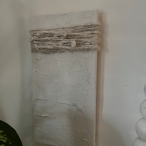 Op de afbeelding: Een wit, getextureerd canvas met een band van natuurlijke vezel touw in het midden. Het canvas is gemonteerd op een witte muur.