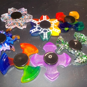 May include: A collection of colorful, flower-shaped fidget spinners. The spinners feature various designs, including rainbow, blue, green, and purple hues. Each spinner has a black center and is made of a clear, possibly resin-like material.
