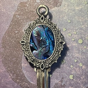 Op de afbeelding: Een zilveren sleutel met een sierlijk ovaal frame met een blauwe en groene abalone schelp inlay.