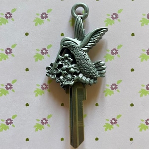 May include: A silver-toned key with a hummingbird and flower design. The hummingbird is perched on a branch with flowers. The key is a house key.