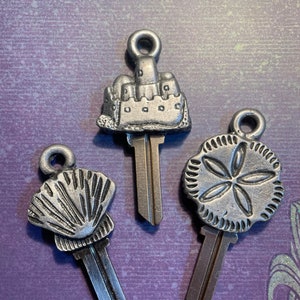 Op de afbeelding: Drie zilveren sleutelhangers met verschillende strandthema's: een zandkasteel, een schelp en een zanddollar. Elke sleutelhanger heeft een zilveren sleutel.