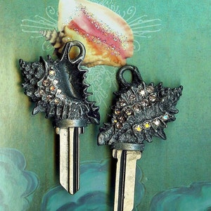 May include: Two silver keychains shaped like seashells with rhinestones. The keychains are on a green background.