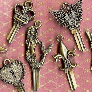 Op de afbeelding: Een verzameling van tien decoratieve sleutelhangers met verschillende ontwerpen, waaronder een zeemeermin, een kroon, een vlinder, een fleur-de-lis en een laars. Elke sleutelhanger is gemaakt van metaal en versierd met strass.