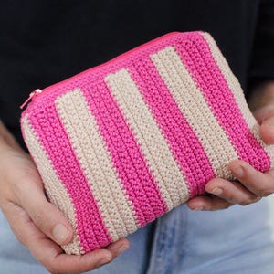 以下が含まれることがあります： ピンクとベージュのストライプ柄のかぎ針編みポーチ。ピンク色のファスナーが付いています。長方形のポーチは両手で持たれており、柔らかく質感のある糸でできています。ストライプは垂直で、等間隔に配置されています。
