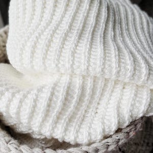 May include: Close-up of a white, ribbed knit hat. The hat is folded over, revealing the textured pattern of the knit. The hat is made of a soft, warm material, perfect for cold weather. The hat is sitting in a basket.
