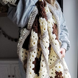 May include: A handmade crochet blanket in a granny square pattern. The blanket features a colour scheme of cream, brown, and tan. The blanket is held up, showcasing the texture and design of the cosy, warm blanket.