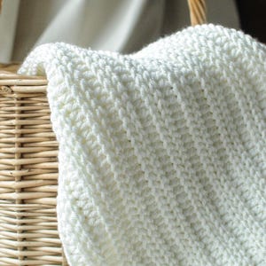 Op de afbeelding: Een wit gehaakt deken in een rieten mand. Het deken is gemaakt met een eenvoudig steekpatroon en heeft een zachte, pluche textuur.