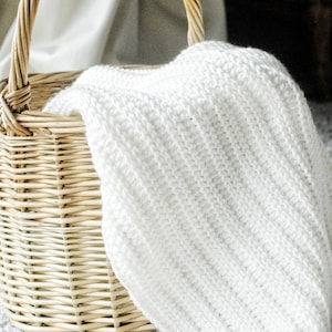 May include: A woven wicker basket with a curved handle holds a folded white knitted blanket. The basket is light brown, and the blanket has a ribbed texture. The scene is set on a light gray carpet.