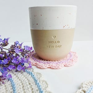Könnte beinhalten: Ein Keramikbecher mit weiß gesprenkeltem Oberteil und mattbeigem Unterteil, der den geprägten Text HELLO NEW DAY und ein kleines Herz aufweist. Der Becher steht auf einem runden, rosa gehäkelten Untersetzer mit Wellenrand neben violetten Blumen.