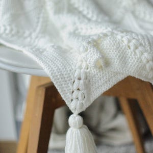 May include: A white crocheted blanket with a detailed pattern and a tassel. The blanket is draped over a wooden chair, highlighting its intricate design and soft texture. The tassel hangs from the corner, adding a decorative element.