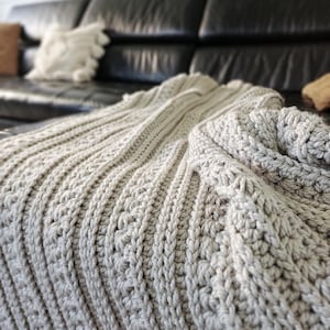 May include: A light gray crochet blanket featuring a textured pattern of vertical ribbed columns and raised stitches. The fabric displays a consistent yarn weight and a dense, looped construction that creates a soft, dimensional surface throughout.