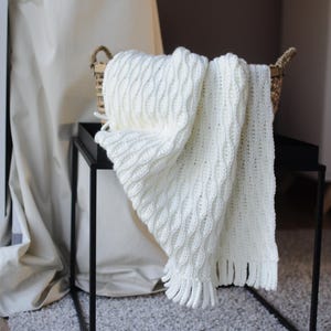 Puede incluir: Una manta blanca de ganchillo con flecos drapeada sobre una mesa auxiliar de metal negro. La manta está en una cesta tejida.