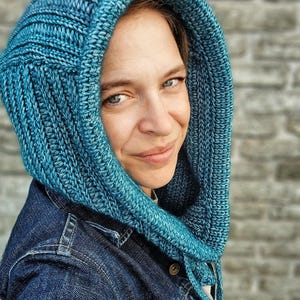 Könnte beinhalten: Eine türkisfarbene Strickhaube mit Rippenstruktur. Die Kapuze wird über einer Jeansjacke getragen. Die Kapuze ist einfarbig und scheint aus weichem Garn gefertigt zu sein. Die Kapuze ist so konzipiert, dass sie Kopf und Hals bedeckt.