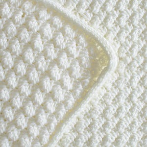 May include: Close-up of a cream-colored crocheted blanket with a textured, raised pattern. The blanket features a rounded corner and a border. The soft, off-white color and detailed stitching suggest a handmade item.
