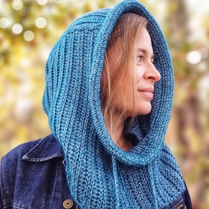 May include: A teal blue knitted hood with a ribbed texture and a thick, rounded edge. The hood features a drawstring closure with matching yarn ties hanging down the front, providing a structured fit around the head and neck area.