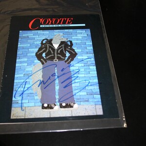 May include: Artwork featuring a cartoon coyote character in a black leather jacket, blue jeans, and sneakers. The artwork is signed and titled "COYOTE A MYTH IN THE MAKING" in red and blue text. The background is a blue brick wall.