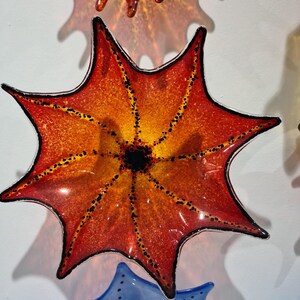 May include: Decorative glass art in the shape of a star, with a gradient of orange and yellow hues. The star has a black outline and is dotted with small black spots. The art is mounted on a white wall.
