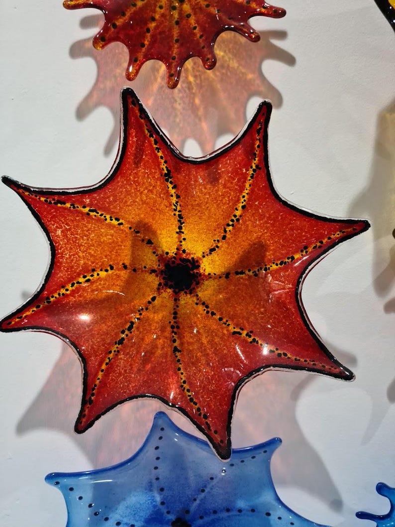 May include: Decorative glass art in the shape of a star, with a gradient of orange and yellow hues. The star has a black outline and is dotted with small black spots. The art is mounted on a white wall.