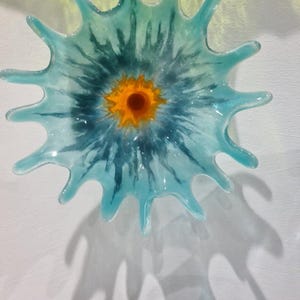 May include: A decorative glass wall art piece in a starburst design. The artwork features a central orange circle surrounded by a blue and teal pattern. The piece casts a shadow on the white wall.
