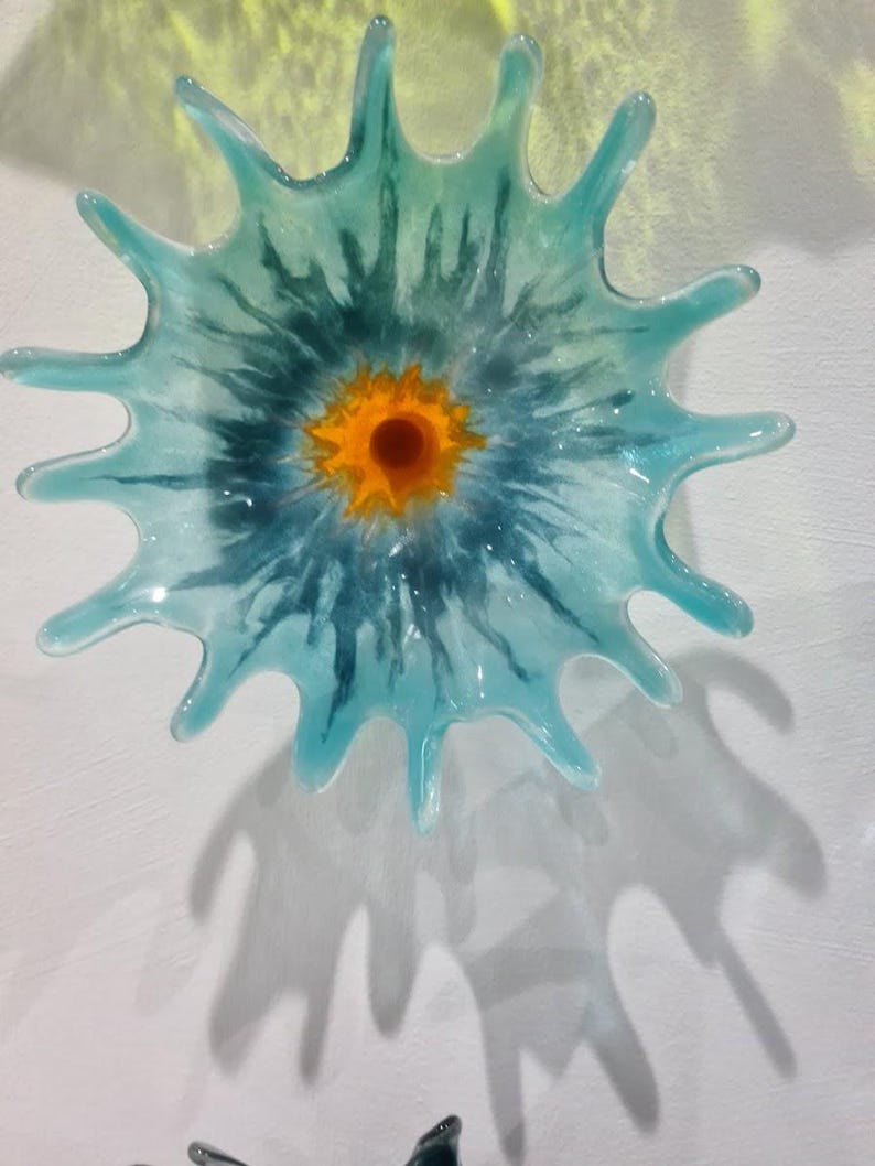 May include: A decorative glass wall art piece in a starburst design. The artwork features a central orange circle surrounded by a blue and teal pattern. The piece casts a shadow on the white wall.