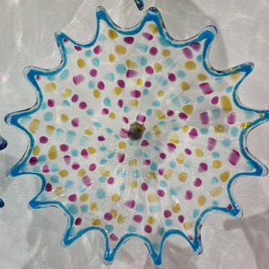 May include: A clear glass decorative plate with a scalloped edge and a blue trim. The plate is decorated with colorful polka dots in yellow, purple, and light blue. The plate is mounted on a white wall.