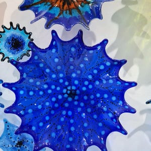 May include: A collection of vibrant, starburst-shaped glass art pieces in shades of blue, orange, and green. The central piece is a deep blue with white polka dots. The glass has a glossy finish, creating a dynamic and eye-catching display.