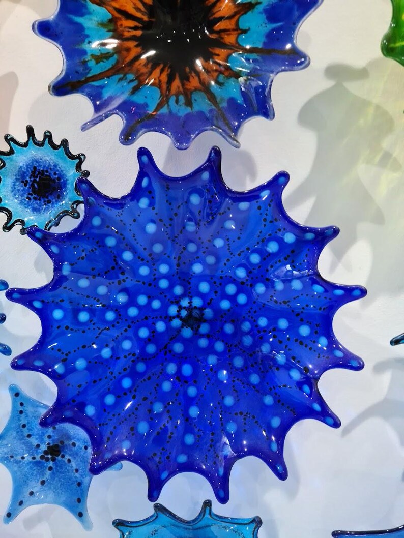 May include: A collection of vibrant, starburst-shaped glass art pieces in shades of blue, orange, and green. The central piece is a deep blue with white polka dots. The glass has a glossy finish, creating a dynamic and eye-catching display.