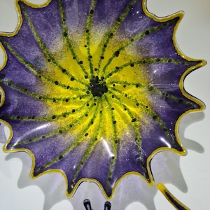 May include: A decorative glass plate with a floral design. The plate features a vibrant color scheme, with a purple outer edge, a yellow center, and green accents. The plate has a scalloped edge and a glossy finish.