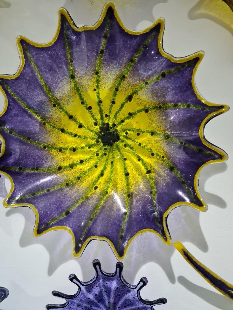 May include: A decorative glass plate with a floral design. The plate features a vibrant color scheme, with a purple outer edge, a yellow center, and green accents. The plate has a scalloped edge and a glossy finish.
