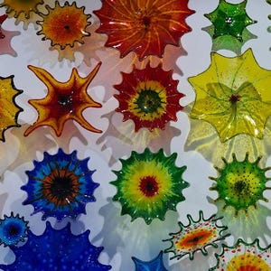 May include: A collection of colorful, spiky glass art pieces in various shapes and sizes. The pieces are in shades of red, orange, yellow, green, and blue, with intricate patterns and textures. The art is mounted on a white surface.