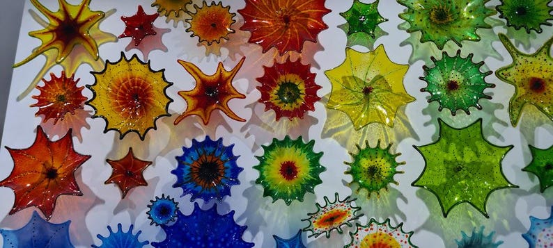 May include: A collection of colorful, spiky glass art pieces in various shapes and sizes. The pieces are in shades of red, orange, yellow, green, and blue, with intricate patterns and textures. The art is mounted on a white surface.