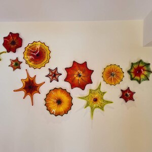 May include: A collection of colorful, glass wall art pieces in various shapes and sizes, including starburst and flower designs. The pieces feature shades of red, orange, yellow, and green, creating a vibrant display on a cream-colored wall.