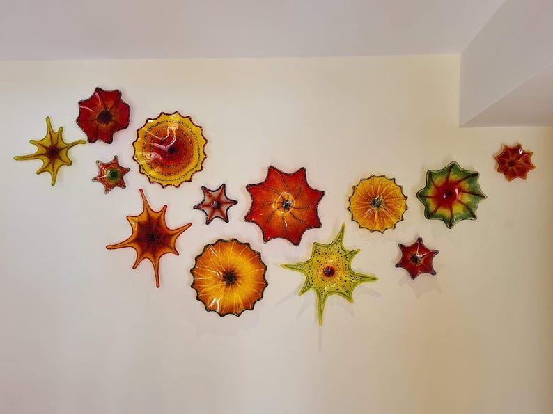 May include: A collection of colorful, glass wall art pieces in various shapes and sizes, including starburst and flower designs. The pieces feature shades of red, orange, yellow, and green, creating a vibrant display on a cream-colored wall.