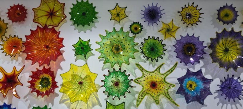 May include: A collection of colorful, spiky glass art pieces in various sizes and hues, including red, orange, yellow, green, and purple. The pieces are mounted on a white wall, creating a vibrant display of decorative art.