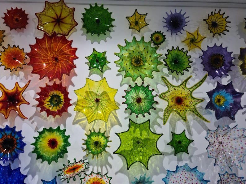 May include: A collection of colorful, spiky glass art pieces in various sizes and hues, including red, yellow, green, and blue. The pieces are mounted on a white wall, creating a vibrant and textured display.